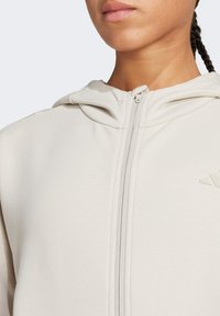 Sudadera con capucha beige claro con cremallera, textura suave, capucha ajustada y detalle sutil del logo en el pecho.