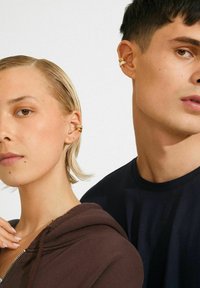 Twee mensen zichtbaar vanaf de schouders omhoog, met gouden oorcuffs, de een in een bruine hoodie en de andere in een zwarte top, tegen een witte achtergrond.