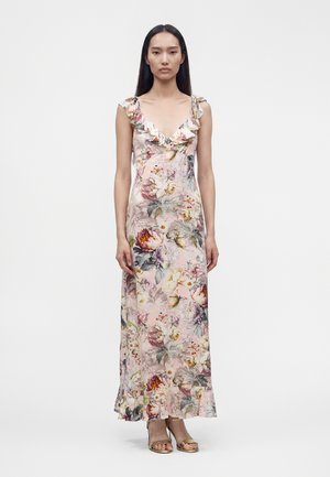 Femme en robe maxi fleurie sans manches avec des volants au niveau du col et de l'ourlet, portant des talons ouverts dorés sur un fond uni.