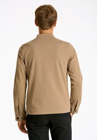 Camisa de manga larga color beige con cuello, puños abotonados, costuras planas y tela suave. Se lleva con pantalones negros. Vista posterior mostrada.