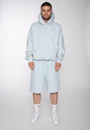 Mann, der frontal steht, trägt ein hellblaues Kapuzensweatshirt und passende Shorts mit weißen Socken und weißen Turnschuhen.
