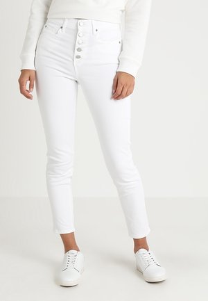 Person wearing high-waisted white skinny jeans with five buttons, paired with white sneakers and a white long-sleeve top.