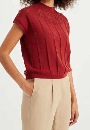 Vrouw die een rood, mouwloos gebreid top met verticale patronen en beige hoge taille broek draagt, hand in de zak, tegen een effen achtergrond.