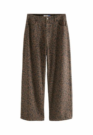 Person, die braune weite Hose mit schwarzem Leopardenmuster und ein schwarzes langärmeliges Shirt vor einem schlichten Hintergrund trägt.