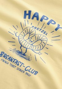 Tessuto giallo con un motivo che presenta un personaggio pretzel sorridente. Il testo recita "HAPPY PRETZEL BREAKFAST-CLUB FRENCH TOAST SERVITO QUOTIDIANAMENTE."