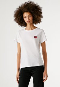 Νεαρή γυναίκα με σγουρά μαλλιά, φορώντας ένα λευκό T-shirt με λογότυπο κόκκινων χειλιών και μαύρο παντελόνι, μπροστά σε ανοιχτό λευκό φόντο.