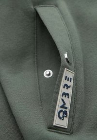 La tela de algodón verde presenta un bolsillo lateral con un botón de presión metálico y una etiqueta cosida con texto azul. Material texturizado y suave.