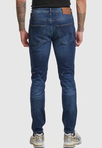 Jeans de mezclilla azul oscuro con corte slim, que cuentan con dos bolsillos traseros y una etiqueta de cuero en la cinturilla. Desgaste sutil cerca de las rodillas.
