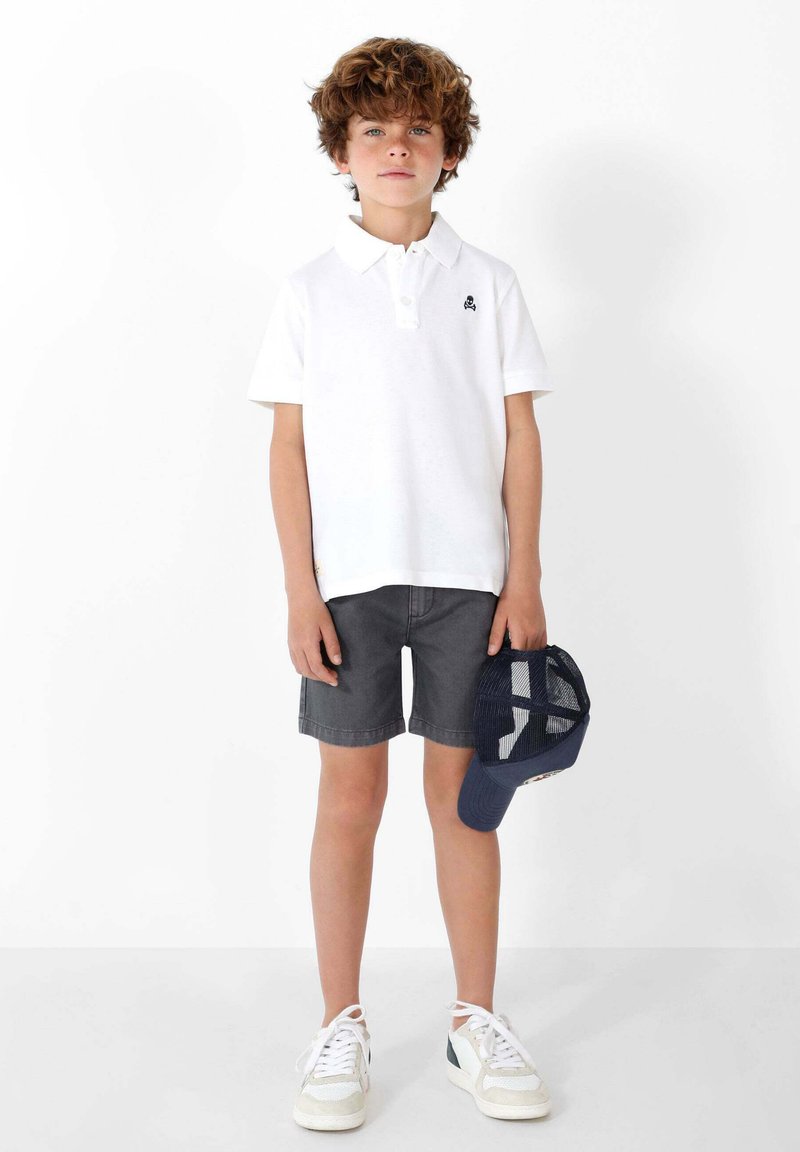 Camisa polo blanca, pantalones cortos grises y zapatillas blancas. Sosteniendo un sombrero azul marino. Tejido suave, ajuste casual, características de diseño mínimas.