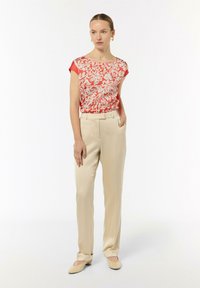 Lekka bluzka w czerwono-białe kwiaty, zestawiona z beżowymi spodniami w kant; modelka stoi z jedną ręką w kieszeni, nosząc płaskie buty.