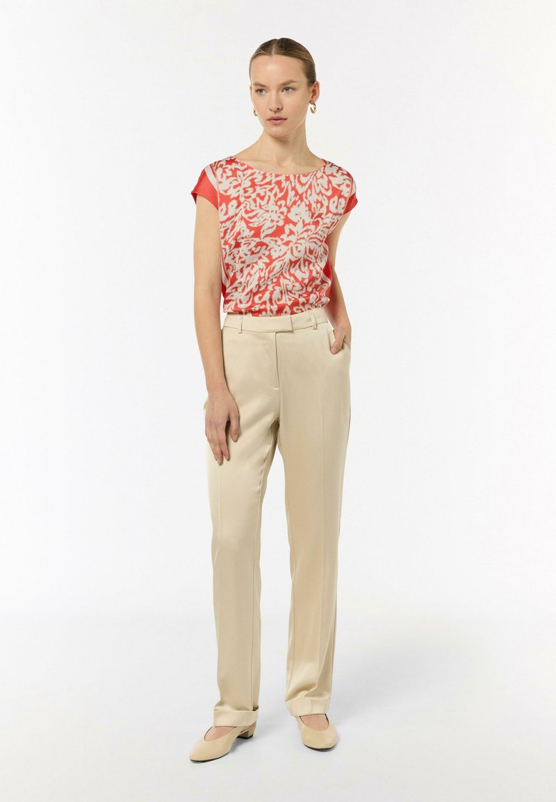 Lekka bluzka w czerwono-białe kwiaty, zestawiona z beżowymi spodniami w kant; modelka stoi z jedną ręką w kieszeni, nosząc płaskie buty.