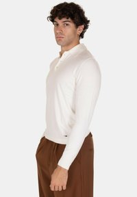Polo shirt bianco a maniche lunghe con colletto, caratterizzato da un'apertura con tre bottoni e un design aderente. Indossato con un pantalone marrone.