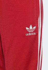 Rdeče atletske hlače iz gladkega materiala z belimi tridelnimi poudarki na straneh in belim Adidasovim logotipom blizu pasu.
