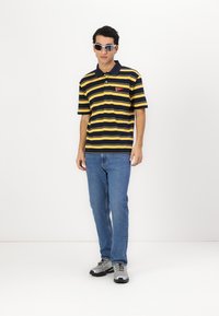 Marineblå og gulstribet polo skjorte med korte ærmer, forsynet med et rødt logo-patch, parret med blå jeans og grå sneakers, samt solbriller.