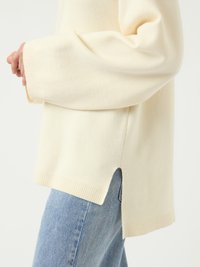 Pull en tricot couleur crème avec des manches larges, des poignets côtelés et des fentes sur les côtés à l'ourlet. Associé à un jean en denim bleu clair.