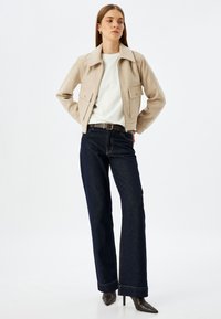 Giacca in misto lana beige con colletto e due tasche frontali, abbinata a jeans in denim a gamba larga blu scuro e stivali neri con tacco.