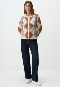 Cardigan en crème, gris et orange brûlé avec un motif argyle ; présentant de grands boutons, des manches longues et une texture douce, associé à un pantalon en denim foncé.