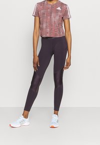T-shirt cropped a fantasia rossa con strisce bianche sulle spalle, abbinata a leggings viola scuro con pannelli lisci e a rete. Sneakers bianche.