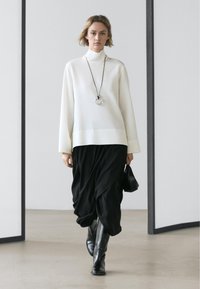 Bílý rolák s dlouhým rukávem, kombinovaný s černými kalhotami s volným padnutím. Modelka má na sobě černé kozačky sahající ke kolenům a náhrdelník s kulatým přívěskem.