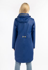 Imperméable bleu avec capuche, taille cintrée et accents en métal doré. Présente un texte imprimé sur le col et un matériau lisse et imperméable.