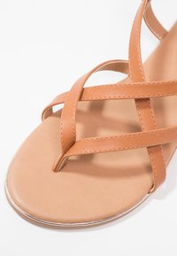 Tan flat sandal featuring thin, crossed leather straps with a smooth sole and toe-post design. Silver trim outlines the sole.
