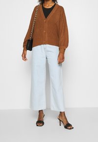 Brun stickad cardigan med V-ringning, knäppning fram och mönstrad textur, ihop med ljusblå vida jeans och svarta klacksandaler.