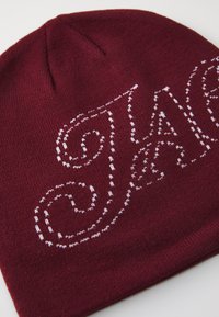 Berretto beanie bordeaux realizzato in tessuto a maglia, con design in testo corsivo bianco ricamato. Tessuto morbido con parte superiore arrotondata.