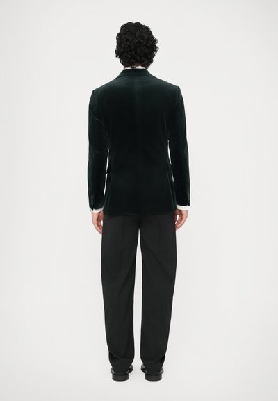 Sametový blazer tmavě zelené barvy s jedním rozventem na zadní straně, v kombinaci s černými kalhotami. Disponuje přizpůsobeným střihem a jemným leskem.