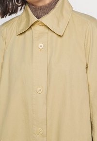 Kvinna som bär en beige knäppt kappa med en bred krage över en brun stickad turtleneck-tröja, med hoopörhängen synliga.