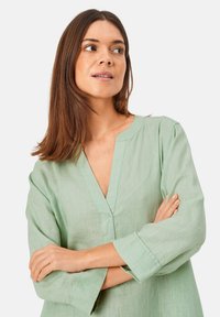 Jasnozielona lniana bluzka z dekoltem w serek, długimi rękawami i luźnym krojem. Posiada delikatną fakturę i przeszycia wzdłuż dekoltu oraz mankietów.