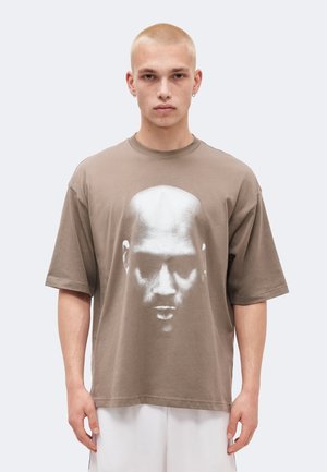 Ung person med barberet hoved iført oversized taupe T-shirt med stort monokromt ansigtsprint, kombineret med hvide shorts.