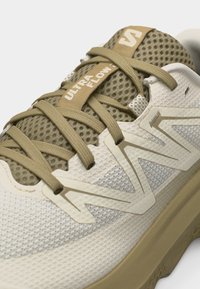 Μπεζ και ελιά αθλητικό παπούτσι New Balance Ultra Flow με επάνω μέρος από δίχτυ, παχιά σόλα και κορδόνια που σχηματίζουν σταυρωτό σχέδιο.