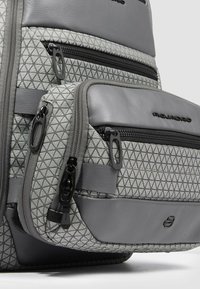 Tasca frontale di uno zaino grigio e decorato con molteplici cerniere nere e logo Piquadro su sezioni di tessuto texturizzato e pelle.