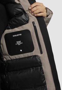 Mustard ja beeži puffer-jakk, millel on tõmblukuga tasku, tekstureeritud nailonist kangas ja must silt, millel on valge tekst ja vahtralehe emblem.