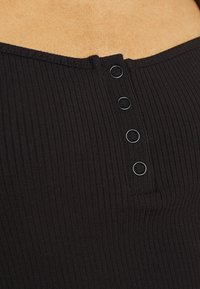 Svart ribbad topp med en rak halsringning och fyra svarta knappar längs mitten. Tyget verkar mjukt med ett strukturerat mönster.