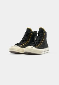 Zapatillas altas negras de lona con acentos amarillos, punta redonda, suela de goma y ojales metálicos para los cordones.
