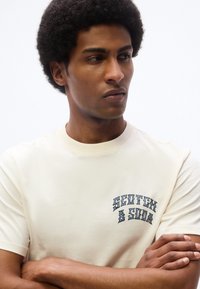 Άντρας με αφρο μαλλιά, φοράει εκρού μπλουζάκι με το κείμενο "Scotch & Soda", με σταυρωμένα τα χέρια, κοιτάζει στο πλάι με φόντο λευκό και απλό.