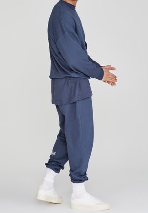 Person in blauem, passendem Sweatshirt und Jogginghose mit weißen Socken und weißen Sneakers, seitlich stehend mit verschränkten Händen.