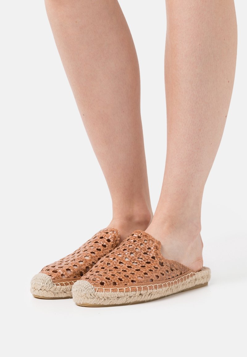 Pieds portant des chaussures espadrilles marron tissées à enfiler avec des semelles en corde tressée sur fond blanc.