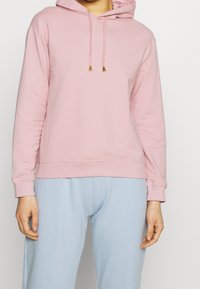 Sudadera con capucha rosa de tela suave, con un bolsillo delantero, cordones ajustables con detalles dorados y puños acanalados. Combinada con pantalones azul claro.