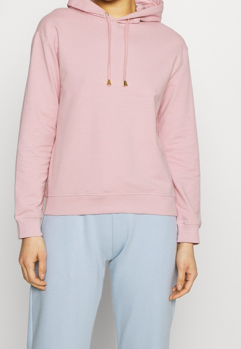 Sudadera con capucha rosa de tela suave, con un bolsillo delantero, cordones ajustables con detalles dorados y puños acanalados. Combinada con pantalones azul claro.