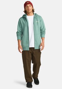 Felpa verde menta con zip e tasche frontali, indossata sopra una maglietta bianca, abbinata a pantaloni marrone scuro e un berretto bordeaux.