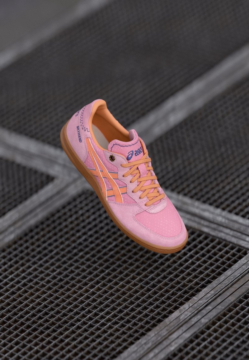 Baskets Asics rose et orange avec semelle en gomme et lacets reposant sur une surface en grille métallique sombre.