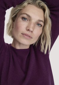 Lila Strickpullover mit weicher Textur, ausgestattet mit einem Rundhalsausschnitt und langen Ärmeln, getragen von einer Person mit kurzen blonden Haaren.