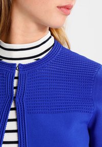 Kvinna som bär en blå texturerad cardigan över en svart och vit randig högkragad topp, visad från hals till axel mot en vit bakgrund.