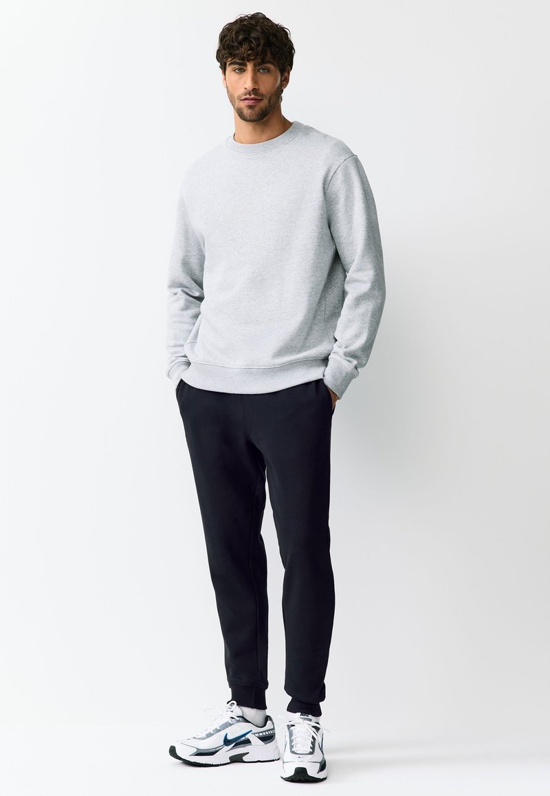 Homme debout, les mains dans les poches, portant un sweat-shirt gris clair, un jogging noir et des baskets blanches avec des touches de bleu.
