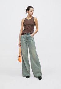 Camiseta sin mangas marrón con un logo, vaqueros anchos de pierna en azul claro y un pequeño bolso naranja. La modelo está de pie con una mano en la cadera. Zapatos negros.