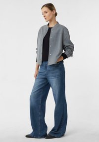 Szara bawełniana kurtka typu bomber z okrągłym dekoltem i zatrzaskami, w zestawie z szerokimi, niebieskimi jeansami o szerokich nogawkach. Czarne buty, jednolite tło.