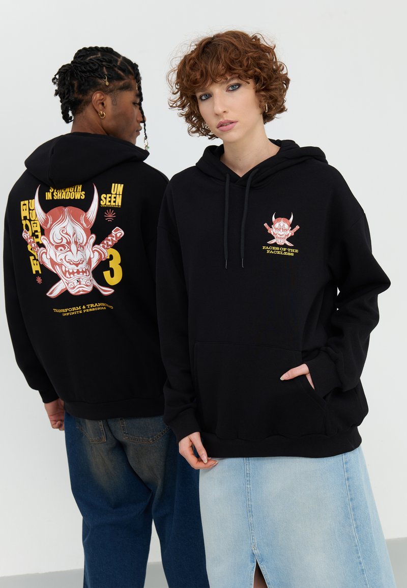 Hoodie noir avec des impressions graphiques au dos et sur la poitrine, présentant un design de masque dans des teintes de rouge et de jaune. Tissu doux, poches avant.