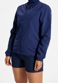 Marineblå zip-up jacka i lätt material med långa ärmar och elastiska manschetter, tillsammans med marinen shorts. Enkelt design med minimala accenter.